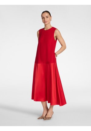 Mujer con un vestido midi rojo sin mangas, con un top ajustado y una falda de satén acampanada, combinado con tacones puntiagudos brillantes y pendientes de aro dorados.