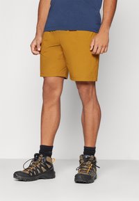 Men's mustard hiking shorts with an elastic waistband, side pockets, and paired with black and orange hiking boots and dark socks.