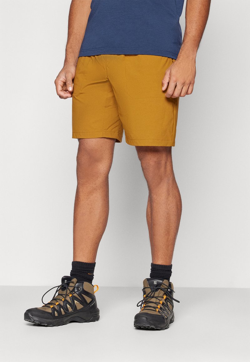 Men's mustard hiking shorts with an elastic waistband, side pockets, and paired with black and orange hiking boots and dark socks.