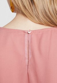 Tessuto rosa con una superficie liscia presenta una chiusura a bottone sul retro, con una leggera fessura sottostante. Orecchini gialli sono visibili nelle vicinanze.