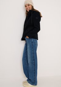 Veste en polaire noire avec capuche, dotée de fermetures éclair et d'un patch, associée à un jean bleu clair à jambes larges et des baskets beiges.