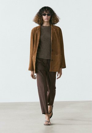 Femme portant une veste en daim marron, un pull marron foncé, un pantalon marron ample, des sandales noires et des lunettes de soleil noires, marchant sur un sol uni devant un mur blanc.