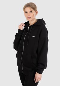 Felpa nera con cappuccio e zip, grandi tasche e tessuto spesso. Presenta coulisse e un piccolo logo sul petto a sinistra.