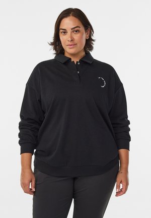 Femme aux cheveux courts et foncés portant un polo noir à manches longues avec un petit texte blanc sur la poitrine, debout devant un fond uni.