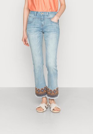Jeans en denim bleu clair avec un bas évasé, présentant un motif géométrique brodé en marron et bleu à l' ourlet, et des bords effilochés.