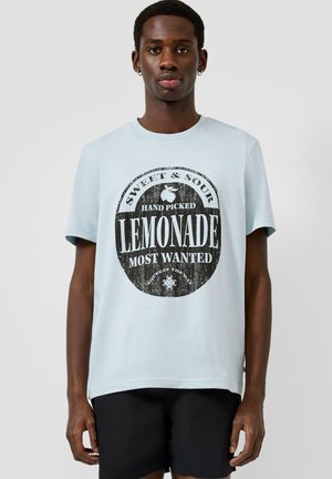 Jonge man draagt een lichtblauw T-shirt met een zwarte cirkelvormige afbeelding 'Sweet & Sour Hand Picked Lemonade Most Wanted', gecombineerd met zwarte korte broek.