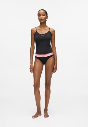 Kobieta stojąca boso, ubrana w czarny top na ramiączkach i czarne majtki z różowym paskiem, na jednolitym białym tle.