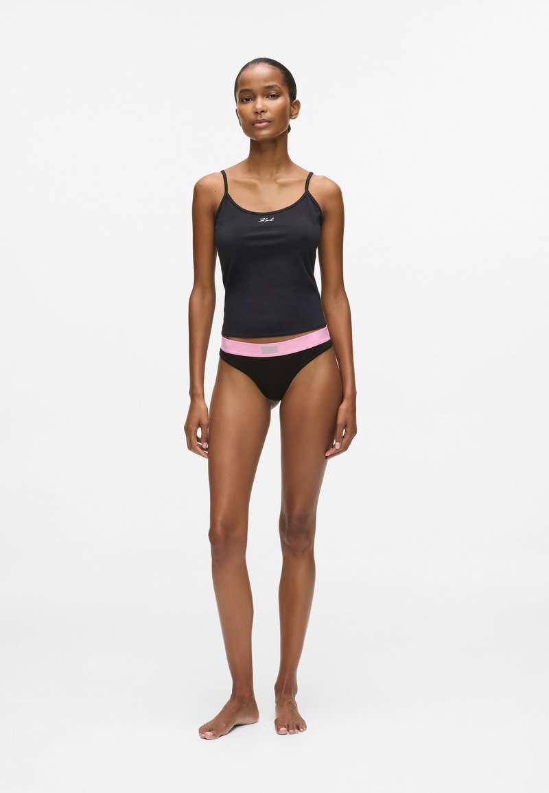 Femme debout pieds nus, portant un débardeur noir et des sous-vêtements noirs avec une ceinture rose, sur un fond blanc uni.