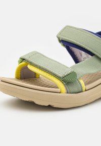 Gröna och gula justerbara remsandaler med en texturerad beige sula, som har en vadderad fotbädd och en blandning av tyg- och gummimaterial.