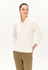 Kermava oversize-polo, jossa on nappilista, kultaiset napit ja kääritty hihansuu, yhdistettynä vaaleanruskeisiin housuihin. Sileä pinta.