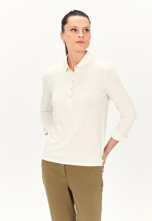 Crème lange mouwen polo-shirt met een knoopsluiting, gouden knopen en opgerolde mouwen, gecombineerd met lichtbruine broek. Zachte textuur.