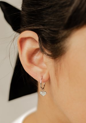 Silver hoop earring featuring a heart-shaped charm adorned with sparkling stones, hanging near the earlobe on a textured, smooth surface.