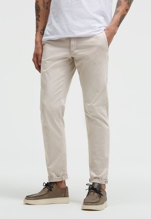 Man wearing beige chinos and brown lace-up shoes, standing with one hand in pocket, visible tattoos on forearms.