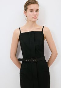 Schwarzer taillierter Jumpsuit mit quadratischem Ausschnitt, dünnen Trägern und tailliertem Gürtel. Verfügt über seitliche Taschen und eine elegante, glatte Textur.