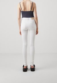 Jean skinny blancs à taille haute ; texture lisse et design ajusté. Associés à des chaussures à plateforme noires, avec des accents frangés.