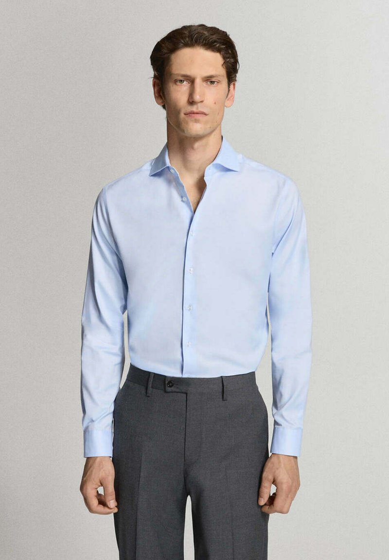 Homme portant une chemise bleu clair boutonnée rentrée dans un pantalon gris foncé, debout devant un fond clair uni.