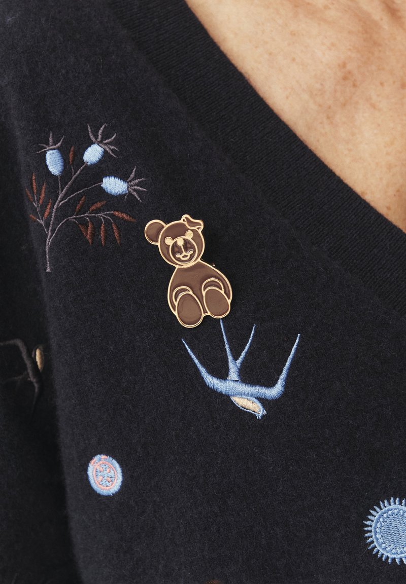 Bronzinė meškos segė su išsamiais detalėmis, prisegta prie tamsios audinio su daugiakalbiu gėlių ir paukščių siuvinėjimu. Minkšta tekstūra.