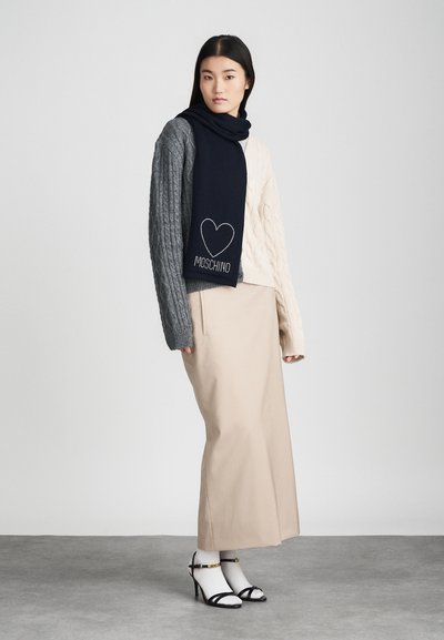 Suéter de malha com seções em cinza e creme, combinado com uma saia longa bege. Um cachecol azul-marinho apresenta um design de coração e "Moschino" em prateado.