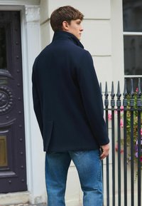 Marineblauer Wollmischmantel mit hohem Kragen, strukturierten Schultern und Seitentaschen, kombiniert mit blauen Denim-Jeans vor neutralem Hintergrund.