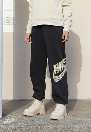 Pantalones de chándal negros con un gran logo de Nike de malla amarilla en el lado izquierdo. Combinados con zapatillas chunky beige, que tienen una suela texturizada.