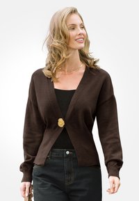 Brązowy sweter z dzianiny, z dekoltem w serek i złotym guzikiem, z długimi rękawami i luźnym krojem, zestawiony z ciemnymi dżinsami.