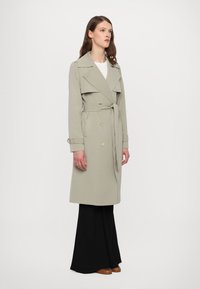 Manteau trench gris clair avec un design croisé, larges revers, ceinture à nouer et accents de boutons dorés, associé à un pantalon noir.