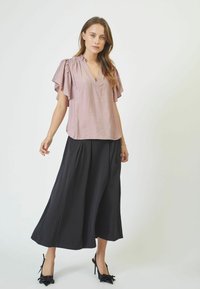 Blusa rosa chiaro con scollo a V e maniche a farfalla, abbinata a una lunga gonna nera. L'outfit è completato da scarpe con tacco alto nere.