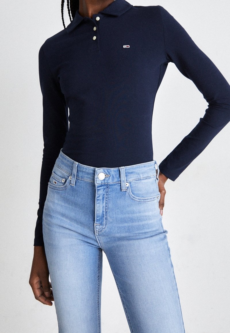 Frau in einem dunkelblauen Longsleeve-Polohemd und hellblauen hochgeschnittenen Jeans, die mit einer Hand auf der Hüfte posiert.