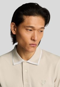 Polo beige con finitura testurizzata, bordi bianchi sul colletto, tre bottoni e un piccolo logo di farfalla ricamato sul lato sinistro del petto.