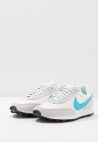 Tenisky Nike s bílým a šedým svrškem, modrým akcentem v podobě swoosh a černou gumovou podrážkou. Hladká kůže a texturovaná semišová materiály.