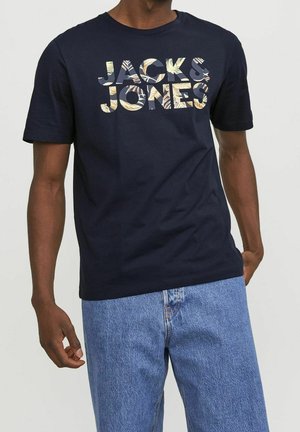 Man wearing a navy blue T-shirt with "JACK & JONES" text in tropical leaf pattern, paired with light blue denim jeans.