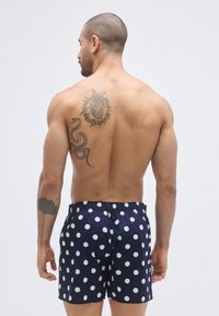 Tengerészkék fürdőnadrág nagy fehér pöttyökkel, rugalmas derékrésszel és bő szabással. Jelentős tetoválások láthatók a háton és a karon.