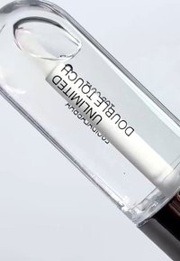 Heldere glazen fles met zwarte dop, gevuld met een heldere vloeistof en een wit etiket met de tekst "UNLIMITED DOUBLE TOUCH."