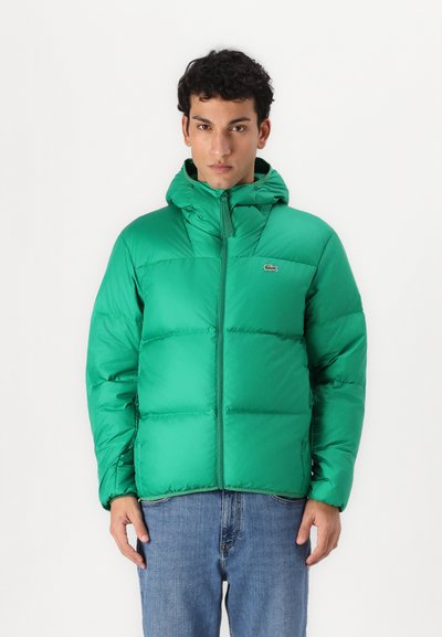 La veste doudoune verte présente une capuche, une fermeture éclair et un design matelassé. Le tissu est lisse avec une finition brillante, assorti à un jean.