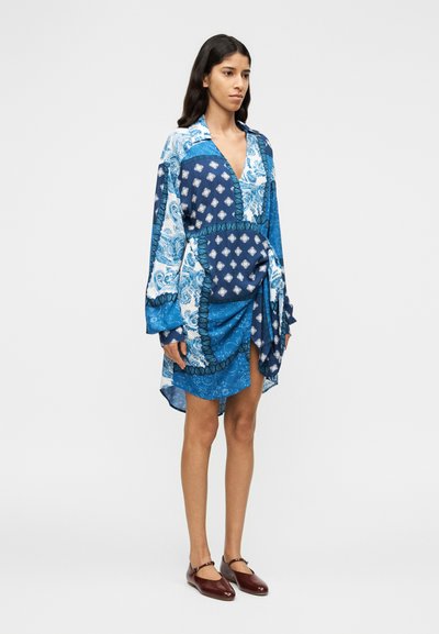 Μπλε φόρεμα τύπου wrap με μακριά μανίκια και σχέδια που συνδυάζουν παϊέτες και γεωμετρικά μοτίβα, με σφιχτή μέση και ασύμμετρο τελείωμα.