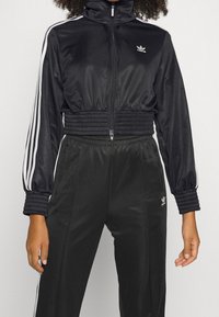 Svart Adidas träningsdräkt med vita ränder på ärmarna och byxorna, elastisk midja och muddar, samt zip-up jacka på en stående person.