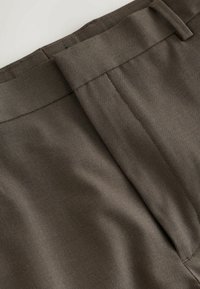 Hnedé nohavice z tkaniny s hladkou textúrou, vybavené plochým predným pásom a jemným záhybom. Šitie je viditeľné pozdĺž okrajov.