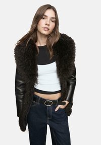 Čierna kožená bunda s hnedým, huňatým kožušinovým golierom, nosená cez čierno-biely crop top. Spárovaná s tmavými riflami s vysokým pásom.