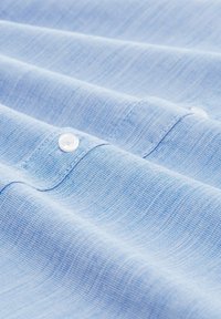 Helles blaues Gewebe mit einer strukturierten Webart, versehen mit einem weißen Knopfdetail. Das Bild zeigt Falten und Knicke im Material.