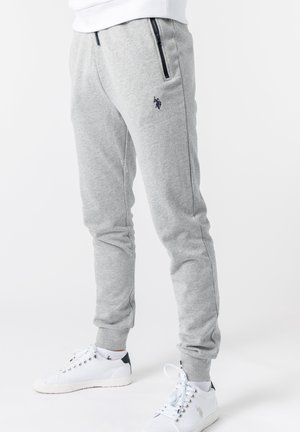 Person trägt hellgraue Jogginghosen mit Reißverschlusstaschen und weiße Sneakers mit schwarzen Fersenlaschen, steht vor weißem Hintergrund.