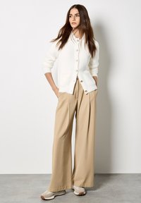 Donna che indossa un cardigan bianco con bottoni, pantaloni beige a gamba larga e sneakers beige, in piedi con le mani in tasca contro una parete neutra.