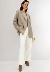 Beige dubbelzijdig jasje over een lichte blouse, gecombineerd met witte gepaste broek en bruine hakschoenen, met een kleine crèmekleurige tas.