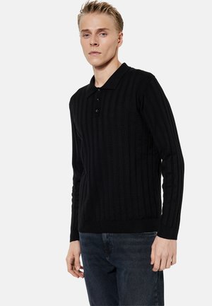 Joven con una camiseta de polo negra de punto acanalado de manga larga y vaqueros oscuros, de pie frente a un fondo claro y liso.