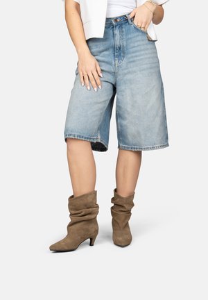 Persoon draagt lichtblauwe denim shorts tot op de knie, een wit shirt, een witte cropped jas en losse bruine suède laarzen met lage hakken.