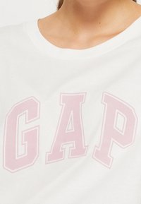 Camiseta de algodón blanca con grandes letras rosas "GAP". El escote es acanalado, con una textura suave y un diseño informal.