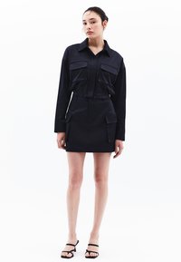 Svart, croppad skjorta med två bröstfickor och matchande högmidjad kjol med sidofickor. Slät tyg, figurnära design, minimal detaljer.