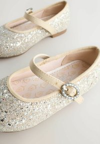 Csillogó balerina cipők ezüst és bézs szövetből, bézs pánttal és dekoratív kör alakú csattal. Simább, lekerekített orrformával.
