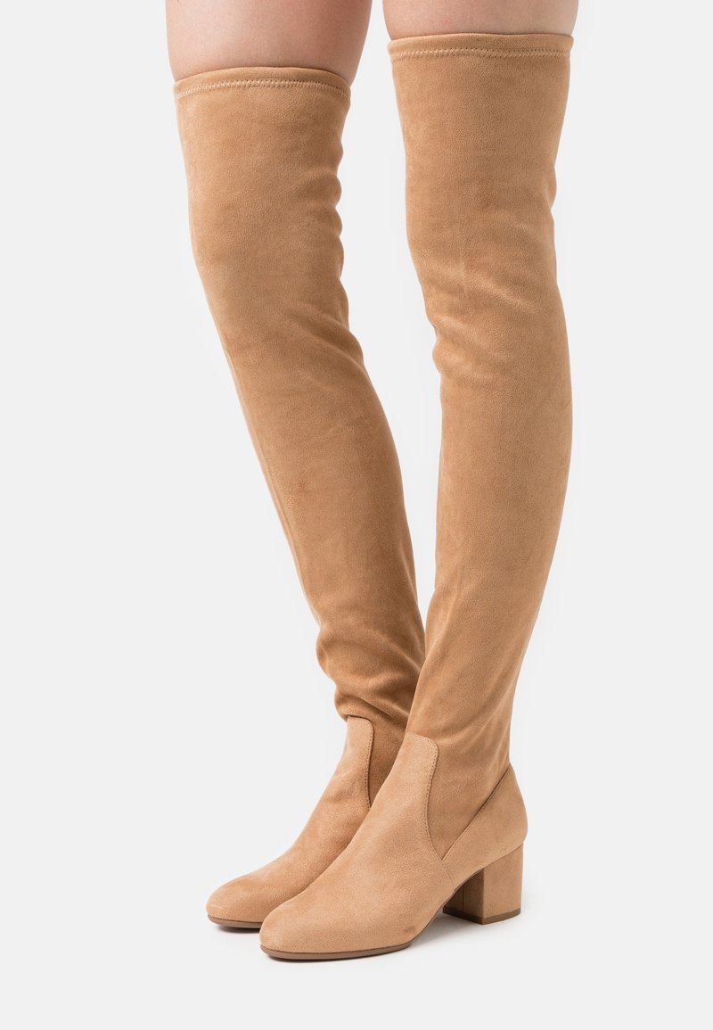 Bottes cuissardes en daim beige avec talons carrés portées sur des jambes sur fond blanc.