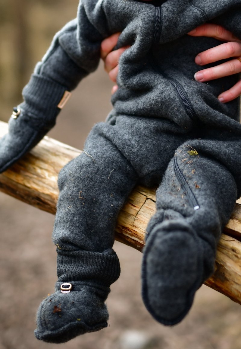 Enfant portant une tenue en polaire gris foncé avec des moufles et des chaussons, assis sur une bûche en bois tenue par des mains d'adultes.