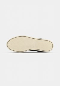 Semelle d'une basket noire montrant une semelle extérieure en caoutchouc beige texturée avec un motif géométrique simple et un bord noir.